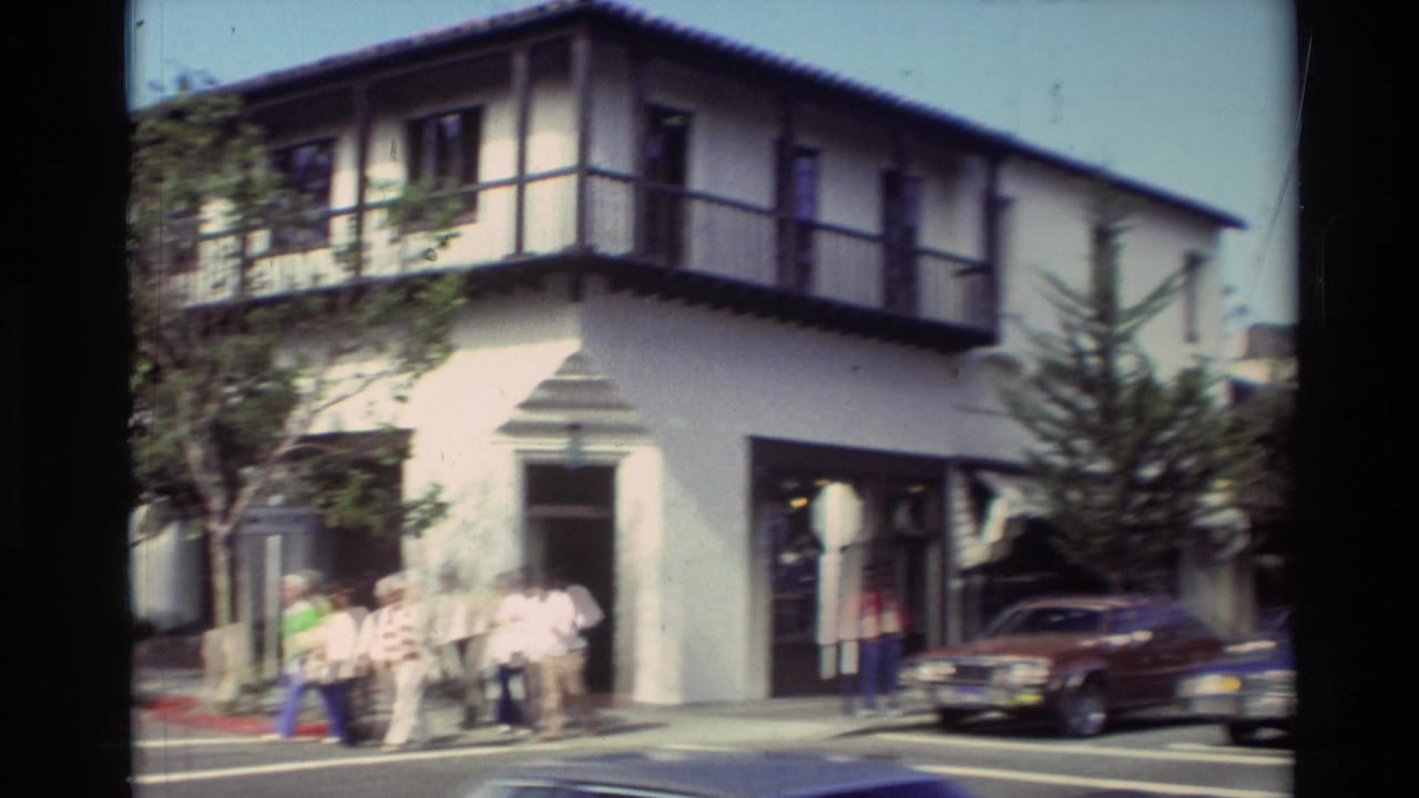 Pedestrians near a white stucco corner building with dark balcony trim and parked cars.