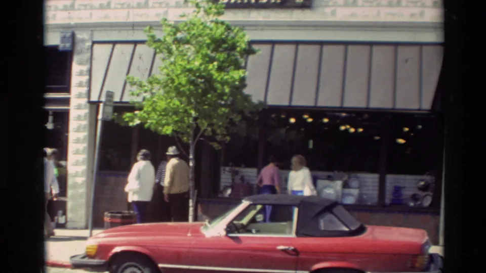 A red convertible and pedestrians outside Carmel storefronts, with awnings and a small street tree visible.