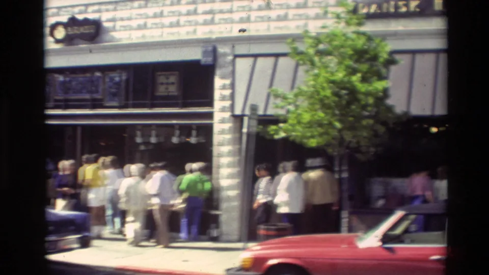 Pedestrians in 1980s clothing gather outside Carmel storefronts as a red convertible is parked in the foreground.