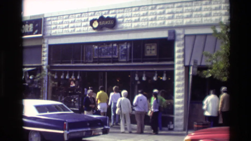 Downtown Carmel storefronts in 1982 with pedestrians on the sidewalk, parked classic cars, and visible shop signage.