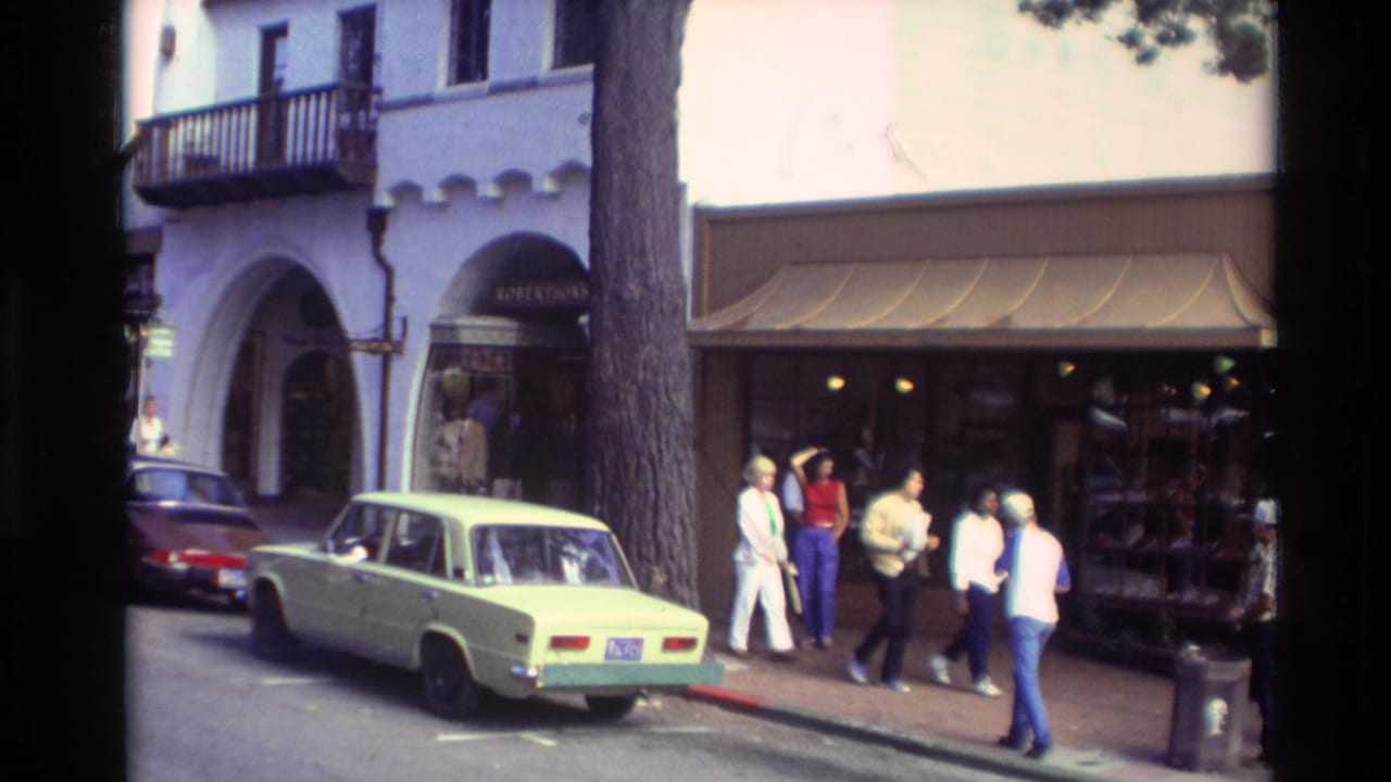 Carmel, California street scene from 1982 with pedestrians near arched storefronts, a parked sedan, and shaded sidewalk displays.