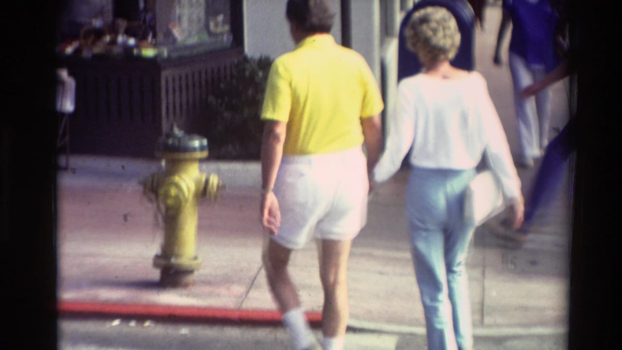 In supported 1982 Carmel footage, a man in a yellow shirt and white shorts walks hand-in-hand with a woman in light blue pants.