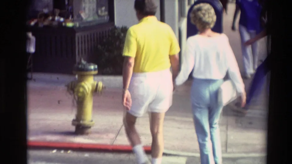In supported 1982 Carmel footage, a man in a yellow shirt and white shorts walks hand-in-hand with a woman in light blue pants.