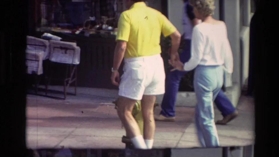 The couple continues down the Carmel sidewalk, filmed from behind with soft 8mm texture and nearby pedestrians in view.