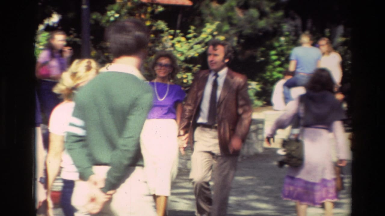 Pedestrians walk on a sunny sidewalk with greenery and a man in a brown jacket approaching.