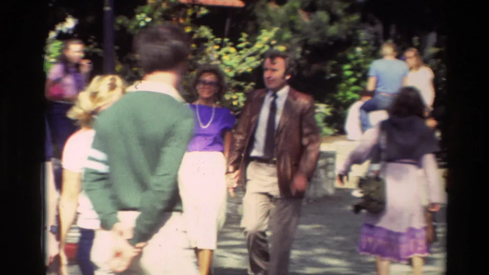 Carmel, California street scene from 1982 with pedestrians in casual 1980s clothing walking past greenery in soft 8mm light.
