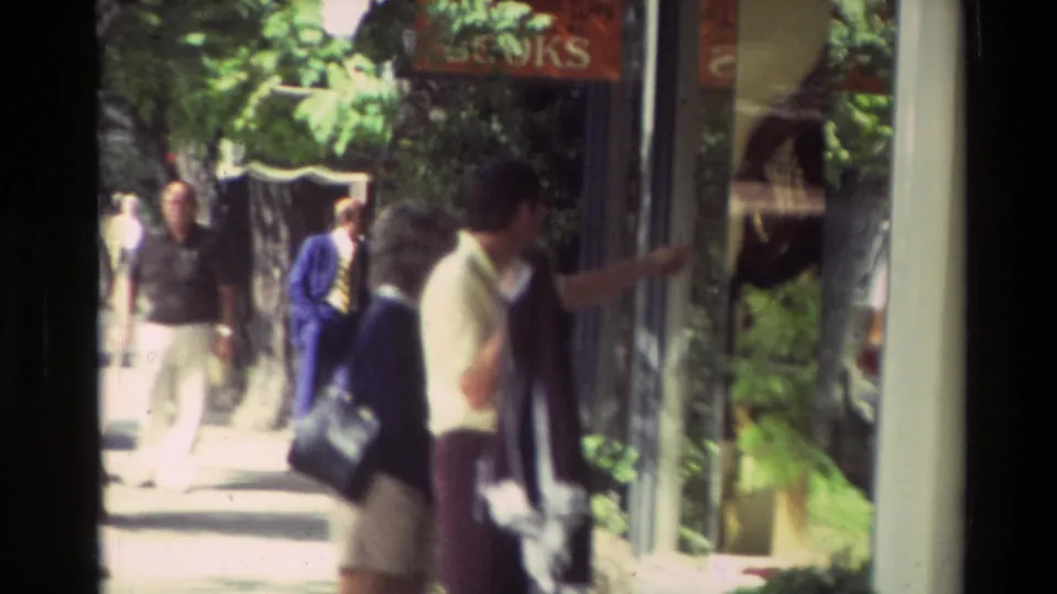 Pedestrians move past storefront windows on a tree-lined Carmel sidewalk, with signage partly visible in the frame.