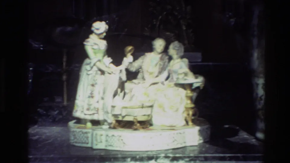 Carmel 1982 home-movie frame showing an ornate porcelain group with standing and seated figures on an oval base behind glass.