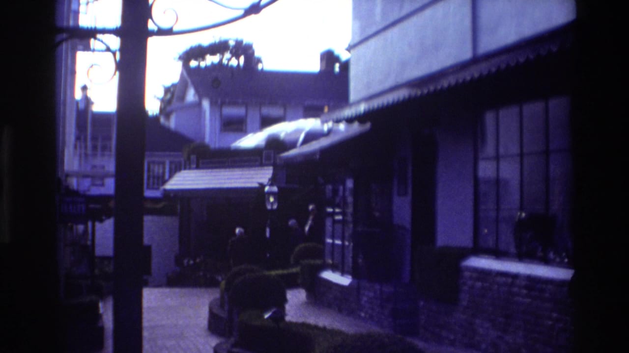 1982 Carmel home-movie frame looking from a shadowed archway into a brick pedestrian courtyard with storefront awnings and trimmed greenery.