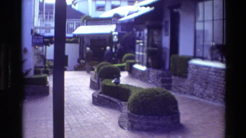 Carmel, California 1982 home-movie frame showing a brick pedestrian lane with landscaping, awnings, and people near the courtyard shops.