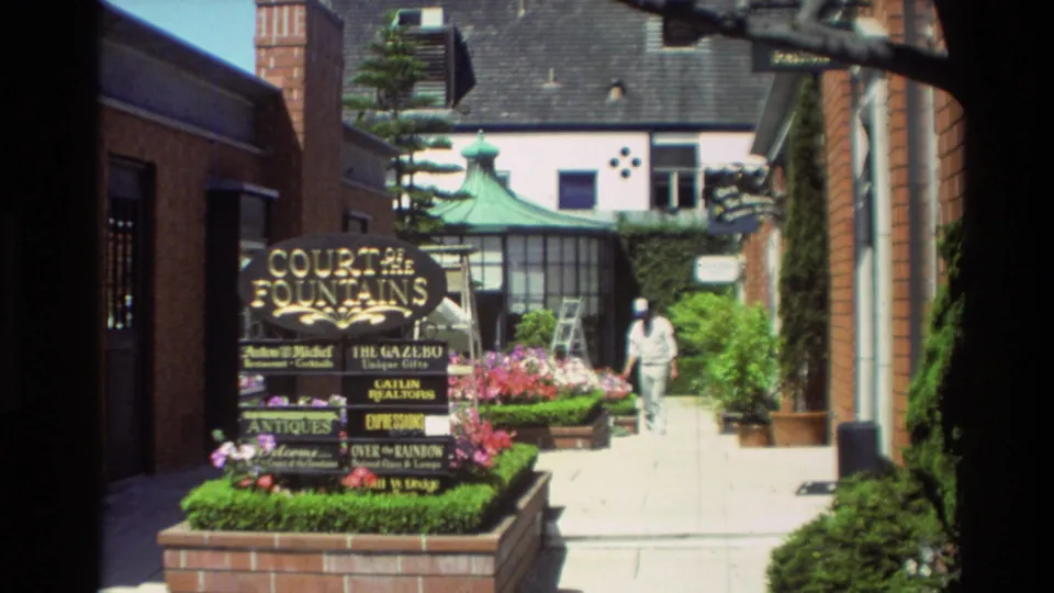 Carmel 1982 frame showing the Court of the Fountains sign, landscaped flowers, brick walls, a green-roofed structure, and a pedestrian walkway.
