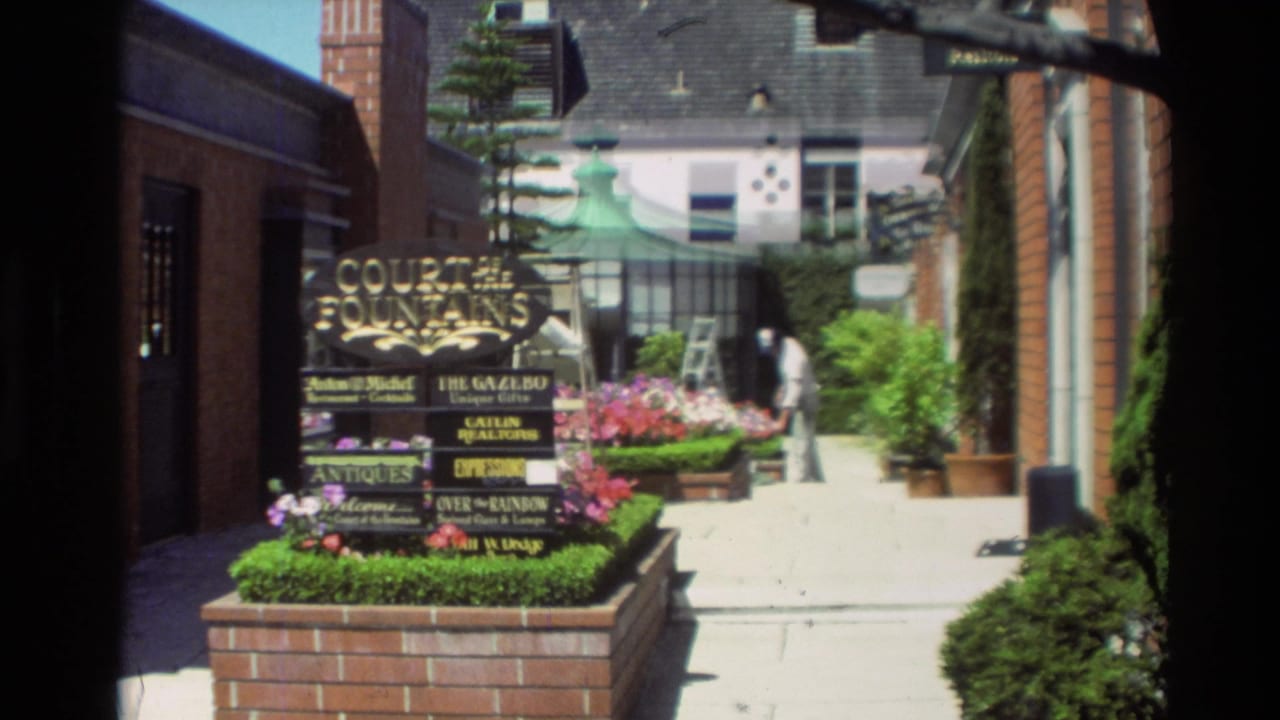 Carmel, California 1982 home-movie frame showing a Court of the Fountains sign, planted brick edging, and a bright walkway between shops.