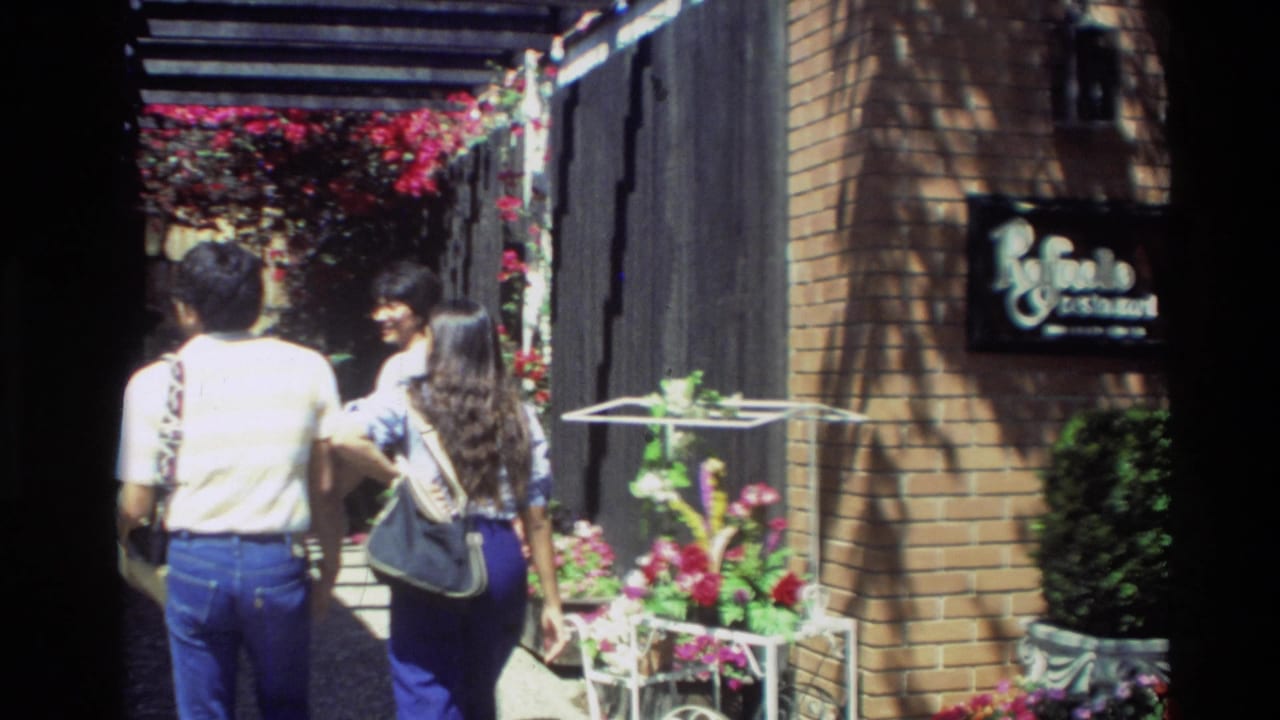 In Carmel, California, two adults in 1980s clothing walk past planters and a storefront sign along a shaded garden alley.