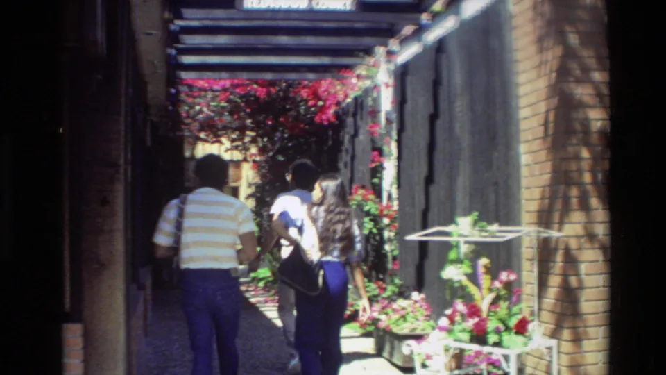 The home-movie camera follows two adults from behind as they walk through a Carmel garden passage with bright flowers and brick storefronts.