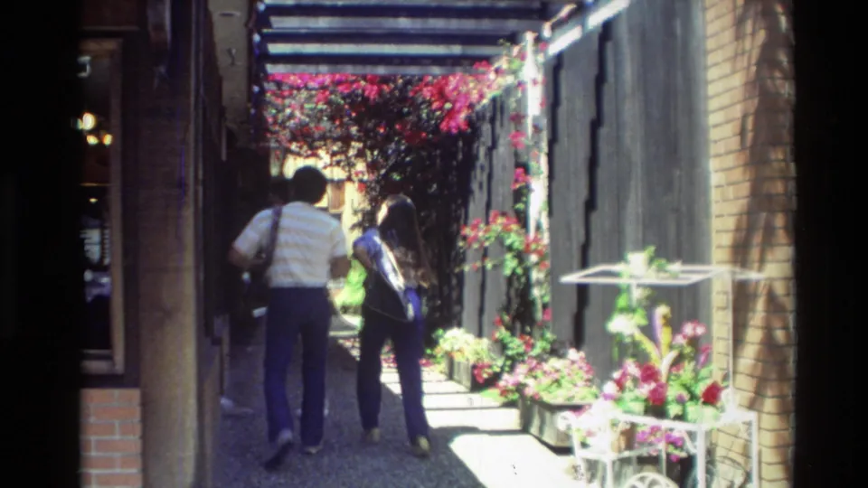 A 1982 Carmel travel frame shows two adults continuing through a sun-dappled alley beside planters, flowers, and shopfronts.
