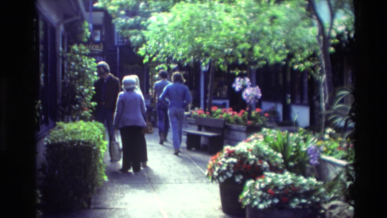 1982 Carmel, California home-movie frame of people walking through a shaded pedestrian walkway with flowers and planters.