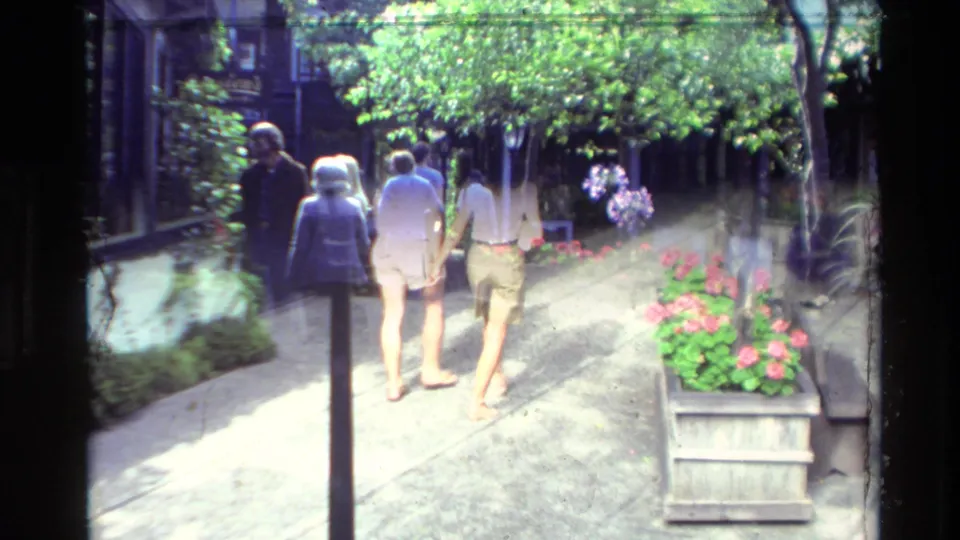 Visitors in casual 1980s clothing walk along a bright Carmel pedestrian path bordered by flowers and planters.