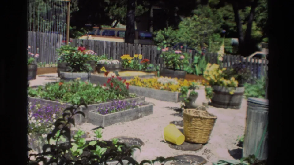 Home-movie frame of Carmel flower beds with gravel paths, planted containers, wicker basket, and yellow watering can, circa 1982.