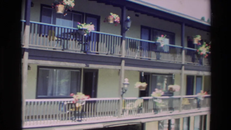 Carmel motel or lodging exterior with two balcony levels, railings, hanging flower baskets, windows, and doors in 1982 footage.