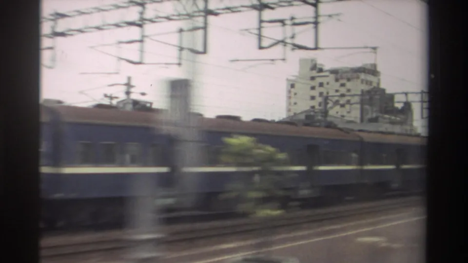 Soft 8mm frame from Taiwan showing blue train cars, overhead rail equipment, and a multi-story building in the background.