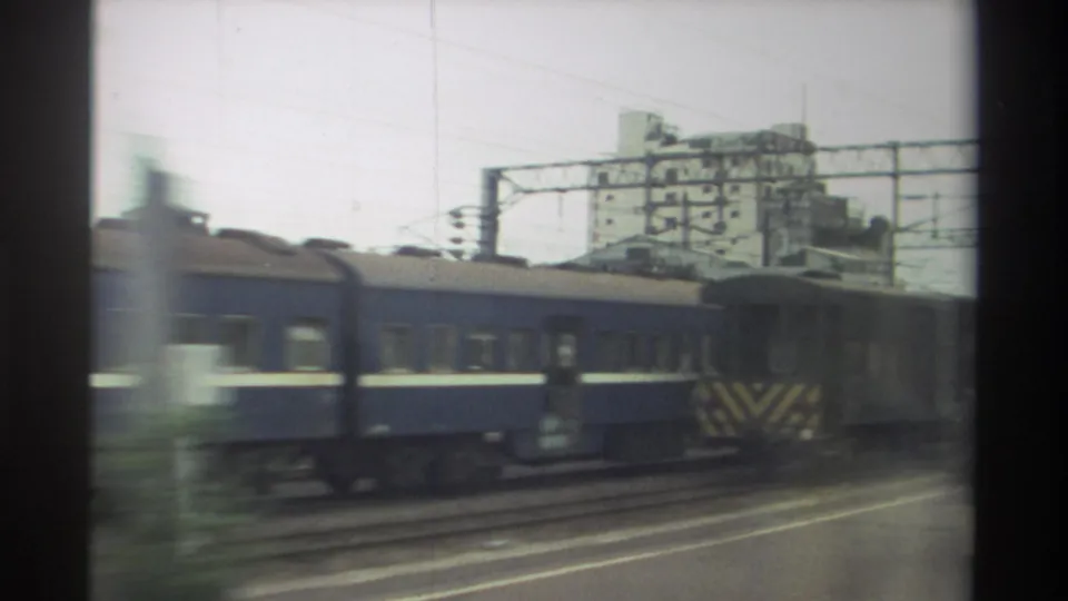1982 Taiwan home-movie frame showing a blue rail car, trackside infrastructure, and a background building from a moving viewpoint.