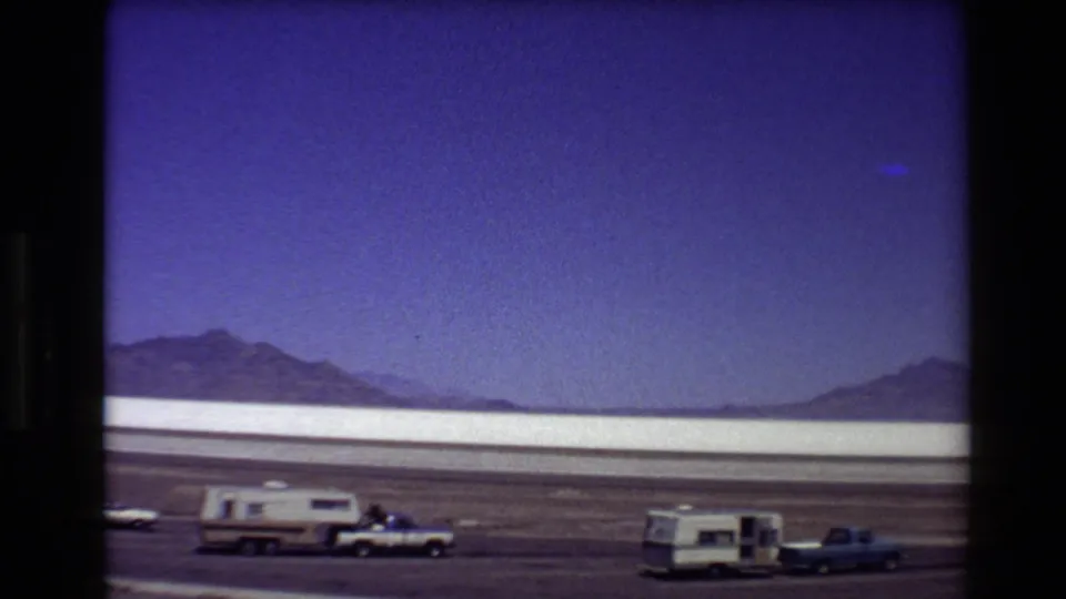 1980 Western USA 8mm frame showing a pickup and camper trailer stopped near a pale salt flat under blue sky.
