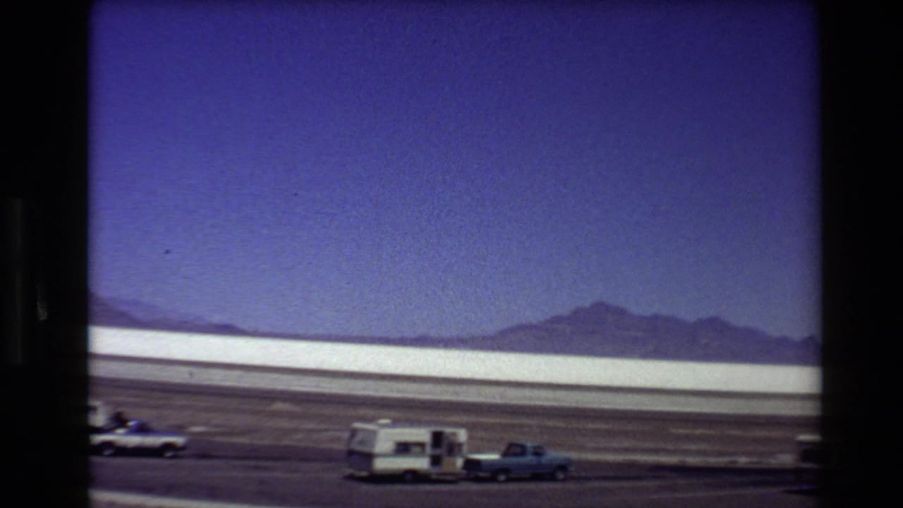 Static 8mm view of a camper trailer and pickup beside a Western USA desert road in 1980.