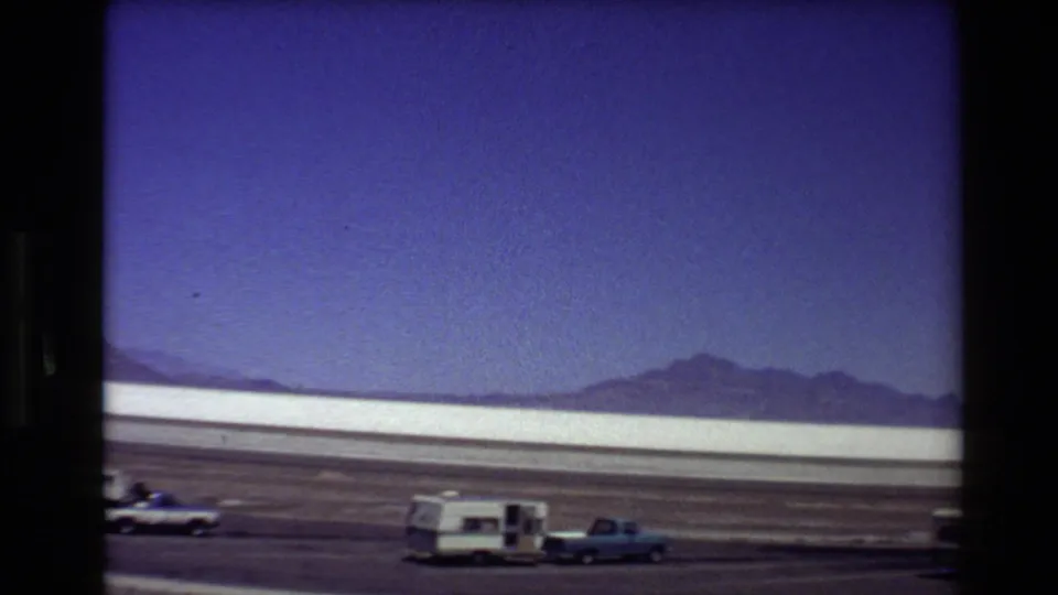 Static 8mm view of a camper trailer and pickup beside a Western USA desert road in 1980.
