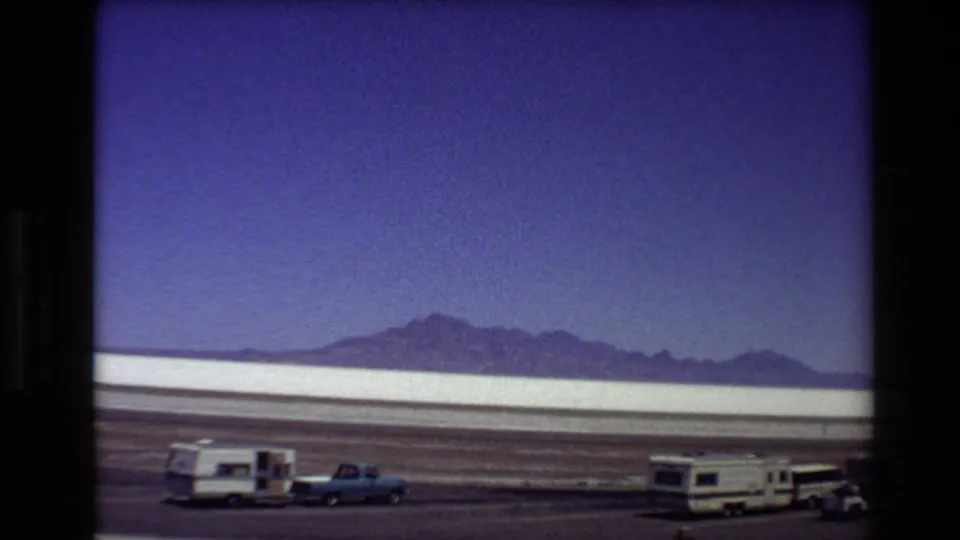 1980 home-movie frame showing two trailer-and-pickup setups along a desert roadside in the Western USA.