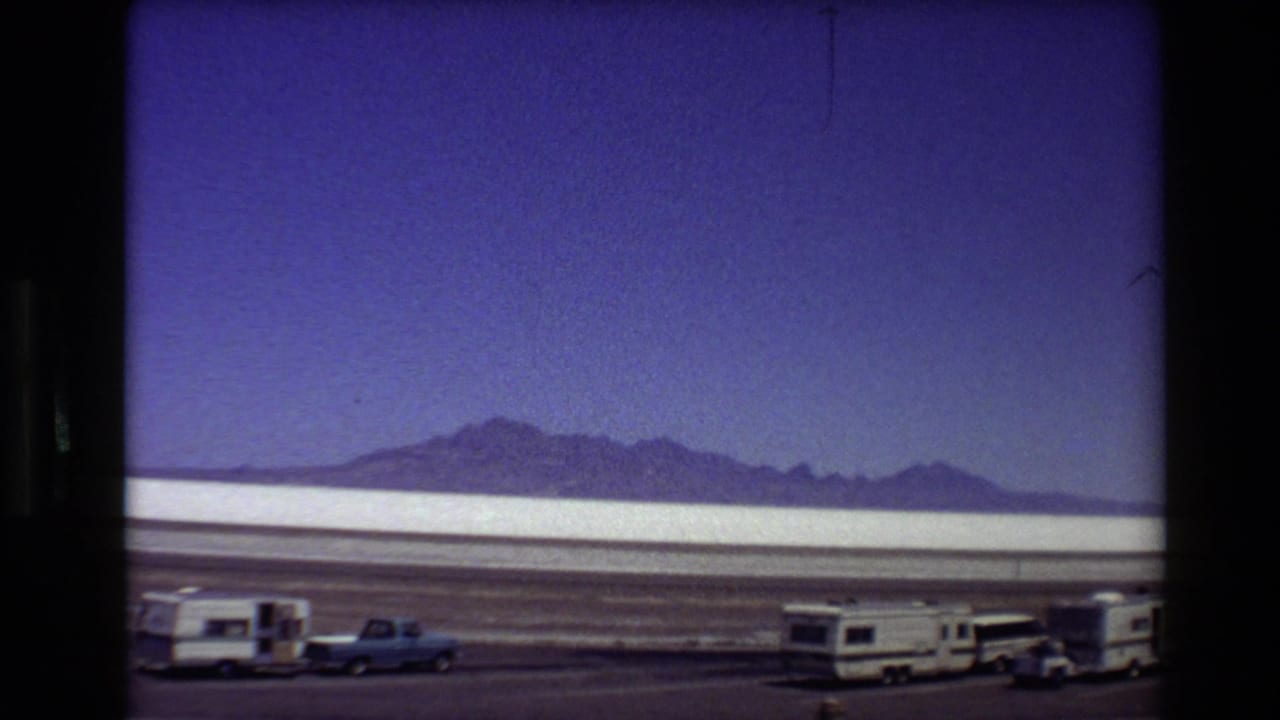 1980 8mm Western USA road-trip view of a salt flat, mountain horizon, parked RVs, and a blue pickup beside a dusty road.