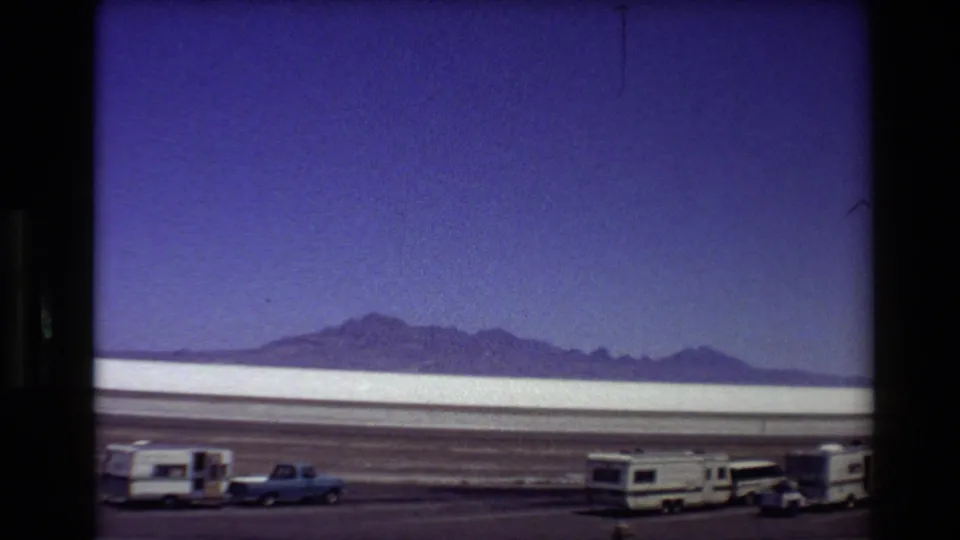 1980 8mm Western USA road-trip view of a salt flat, mountain horizon, parked RVs, and a blue pickup beside a dusty road.