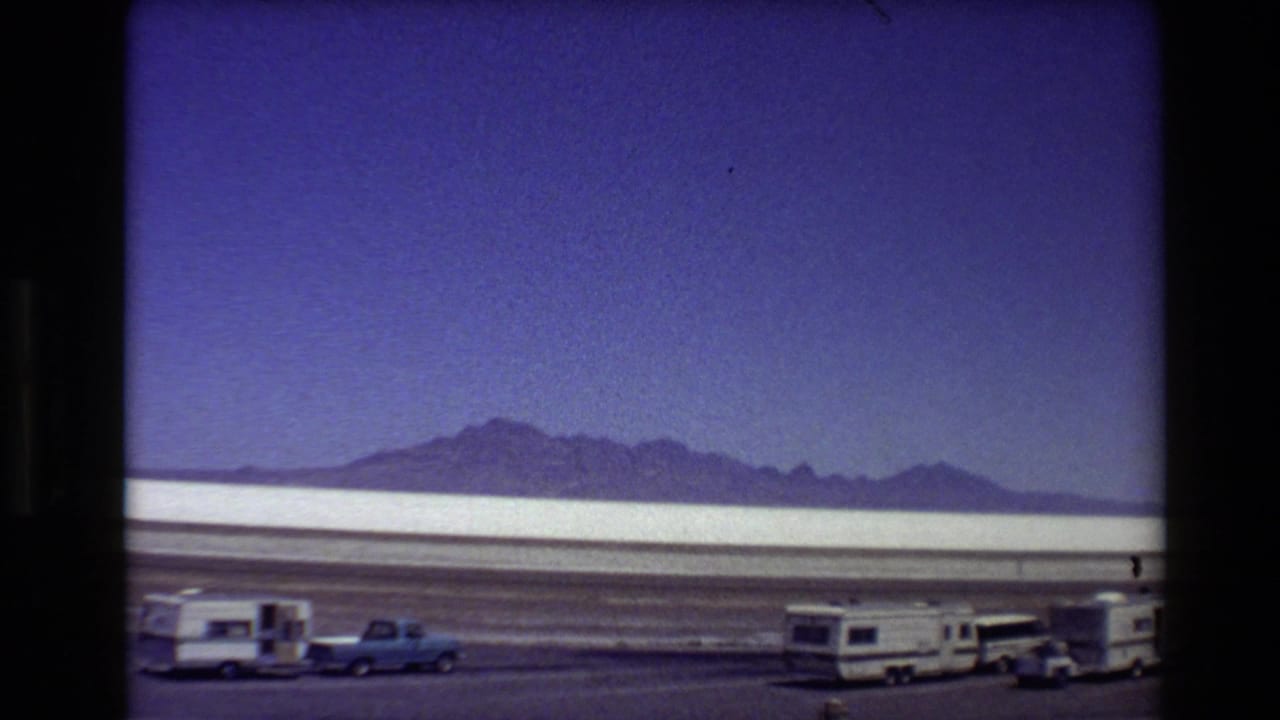 Roadside 8mm view across a Western USA salt flat with RVs, a blue pickup, and mountain ridges on the horizon.