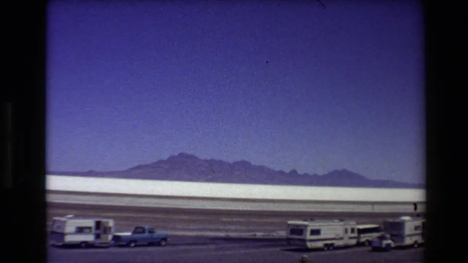 Roadside 8mm view across a Western USA salt flat with RVs, a blue pickup, and mountain ridges on the horizon.