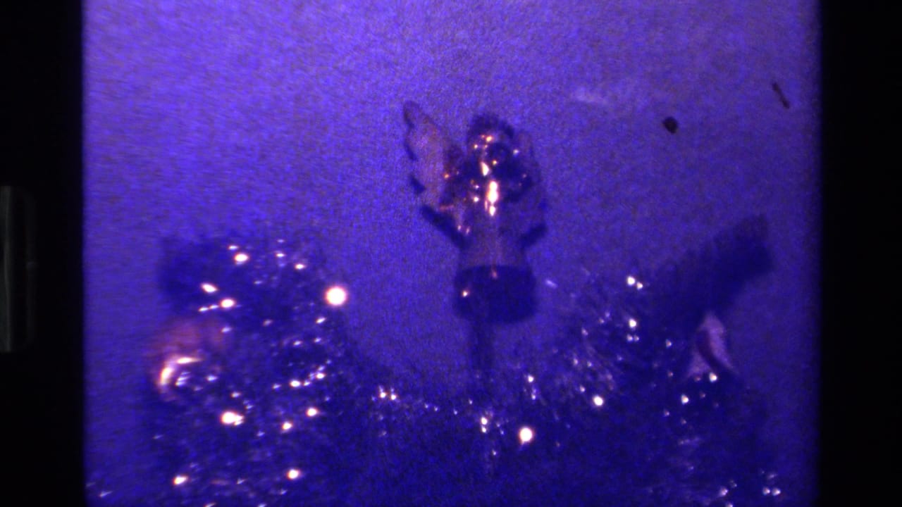 Close-up of Christmas tree tinsel, lights, and a small ornament against a purple background.