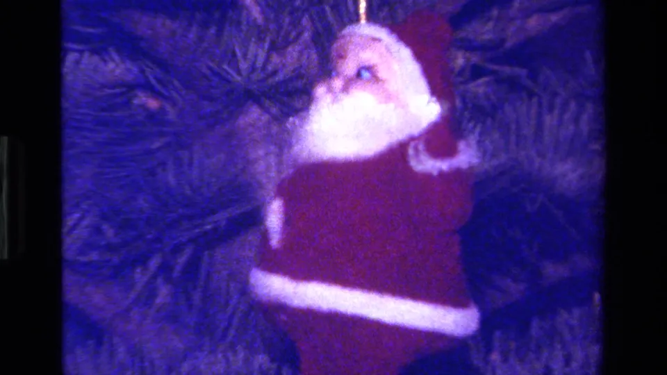 Late-1970s 8mm frame of a Santa ornament in profile, hanging from a Christmas tree in blue-shifted home-movie color.