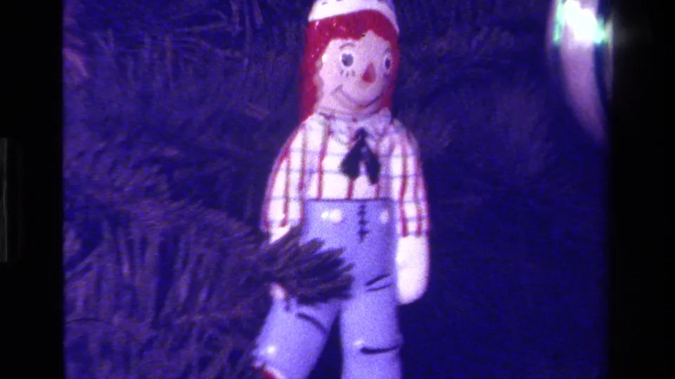 1979 California 8mm frame with a Raggedy-style cloth doll ornament in a checkered shirt and blue overalls on a dark Christmas tree.