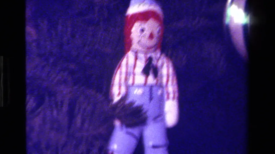 1979 California 8mm frame showing a softly focused cloth doll ornament in overalls against dark evergreen branches.
