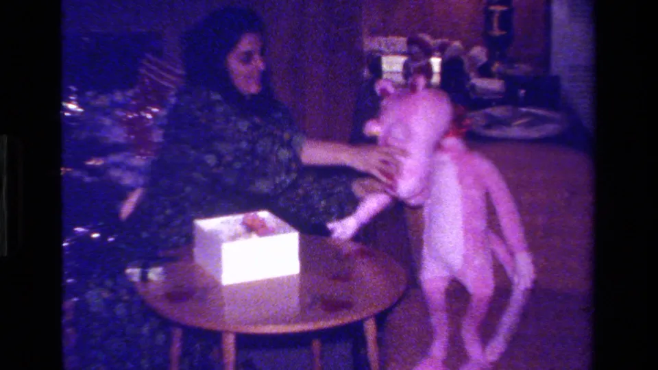 In a 1979 California home-movie frame, a woman in a patterned dress adjusts a large pink stuffed animal beside a small table.
