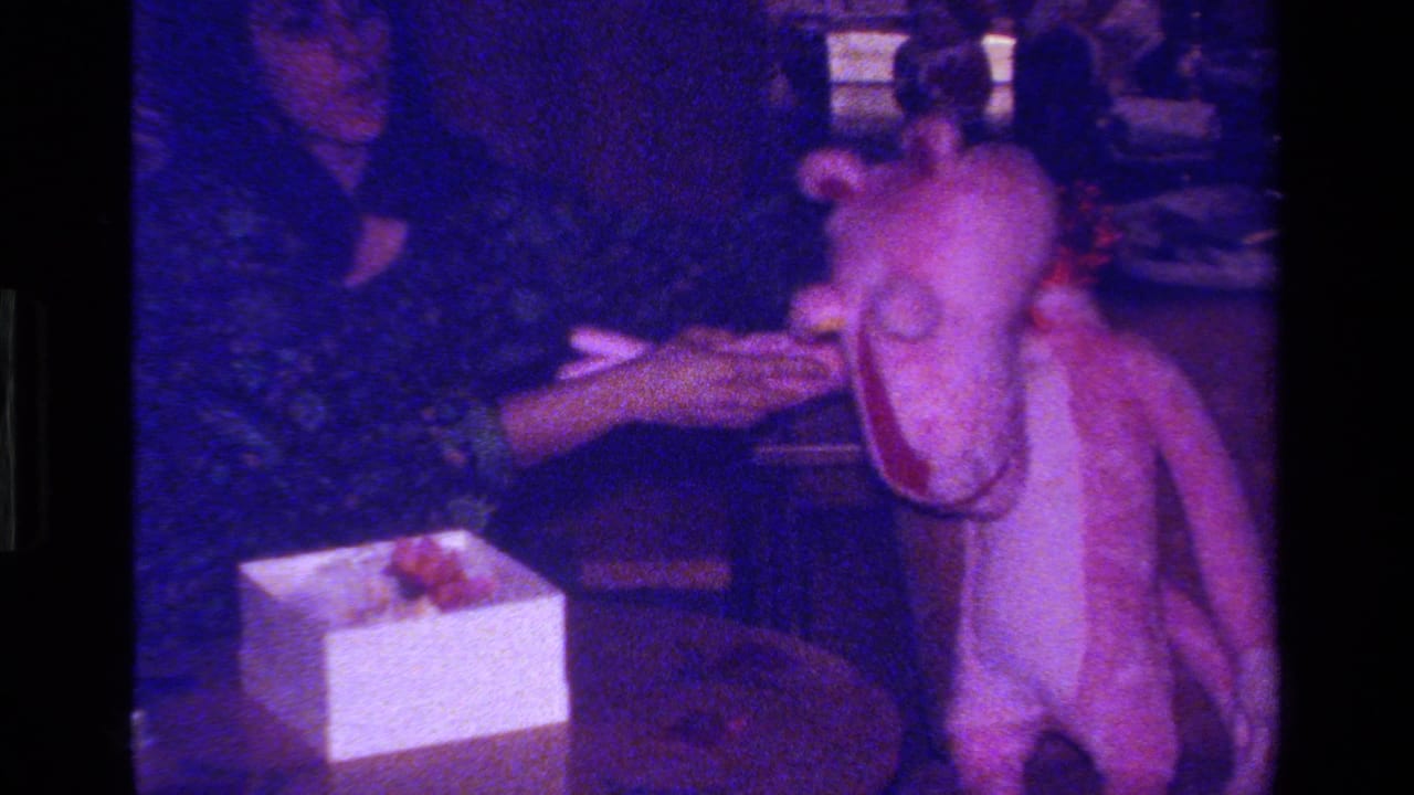 The woman steadies a large pink stuffed animal near a white gift box and small table in the color-shifted 8mm scene.