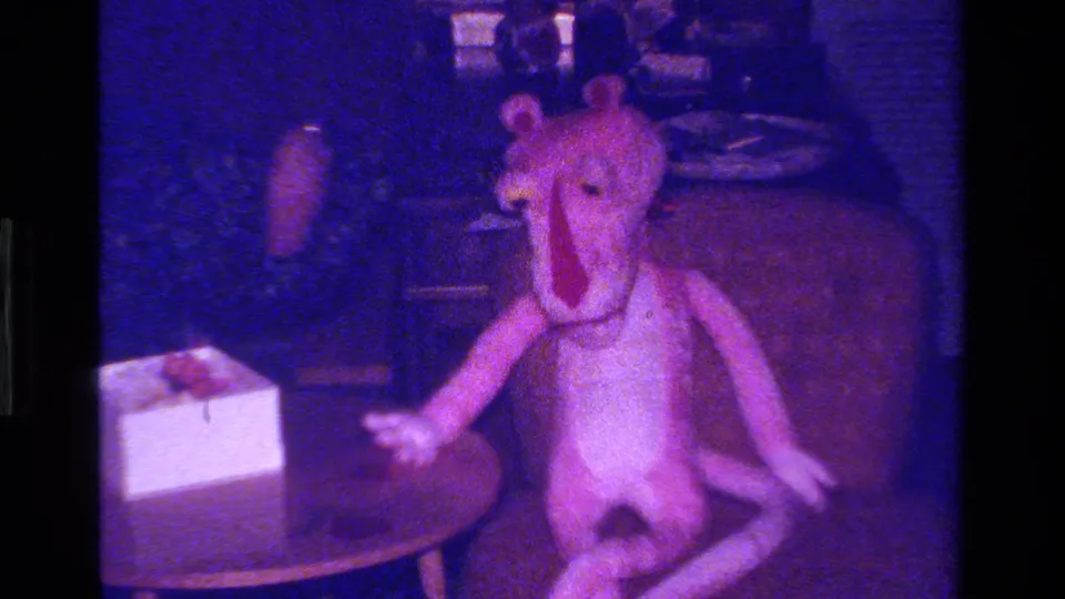 A large pink stuffed animal is positioned beside a small round table with a white gift box in a dim private interior.