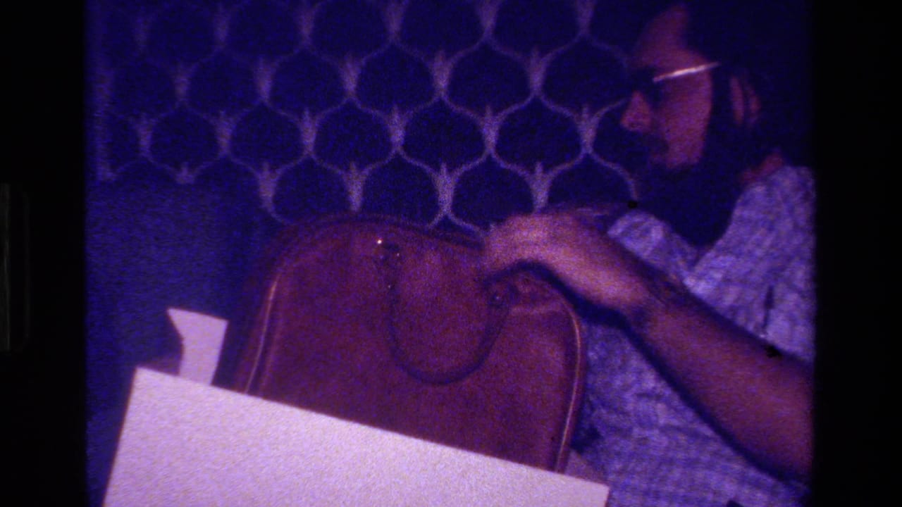 Bearded man with glasses sits beside a brown leather briefcase in a dim room.