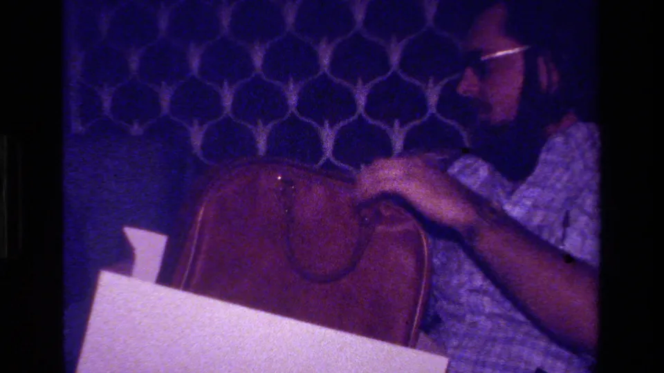 In 1979 California home-movie footage, a bearded man with glasses sits near a brown leather briefcase against blue geometric wall decor.