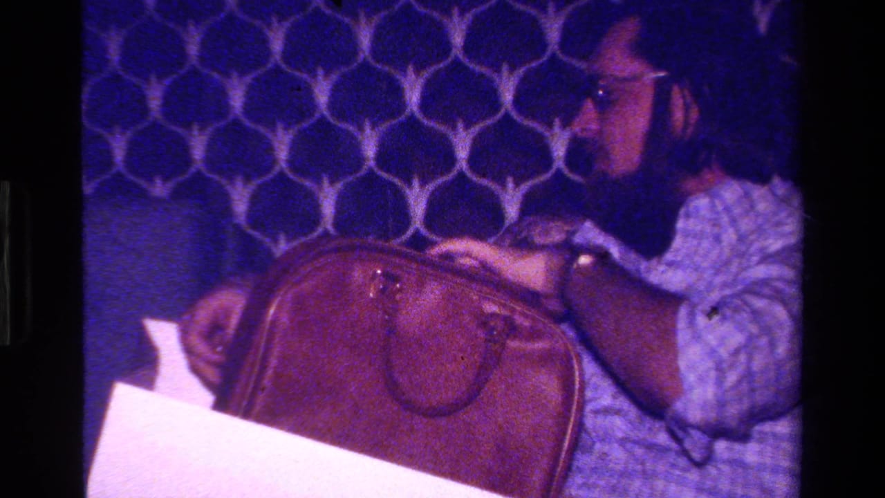 Man with glasses reaches toward a brown leather briefcase on his lap or table.