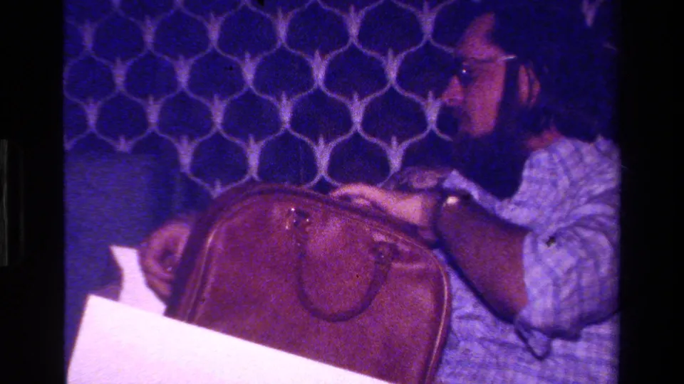 The man appears to inspect or open a brown leather briefcase, with blue patterned wall decor visible behind him.