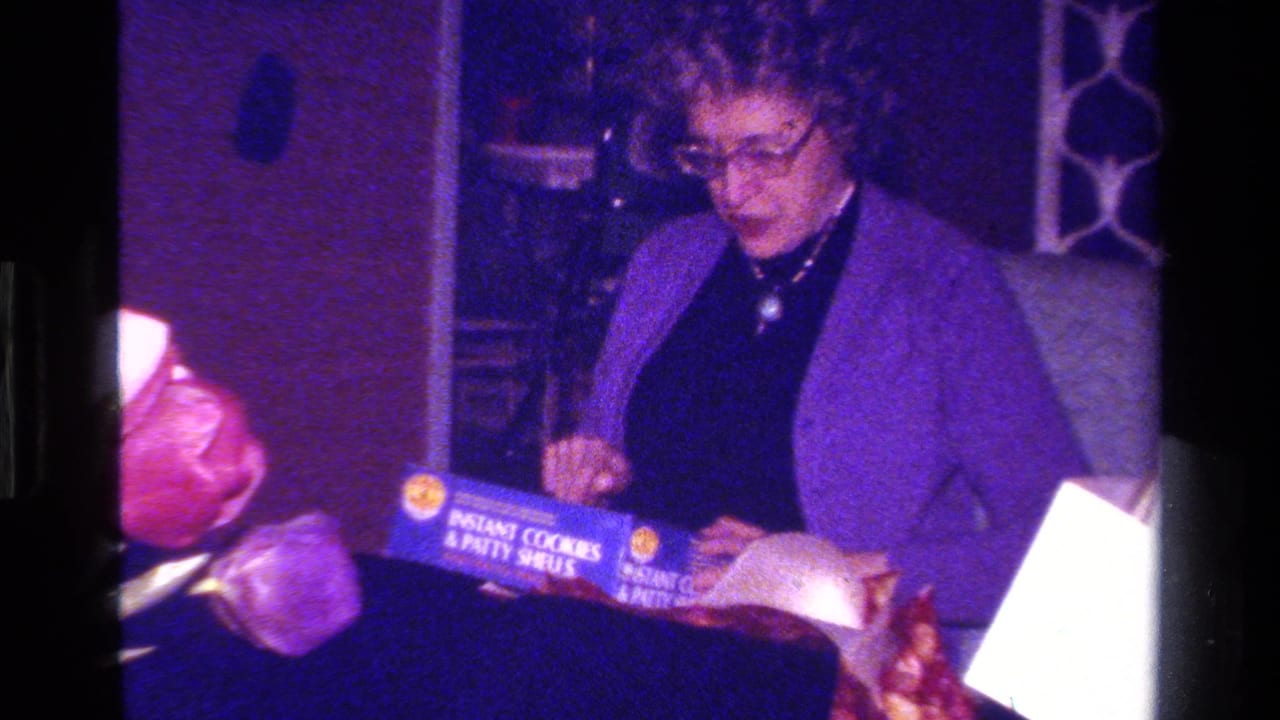A woman in glasses and a purple jacket holds a box labeled as instant cookies and patty shells in a private interior.