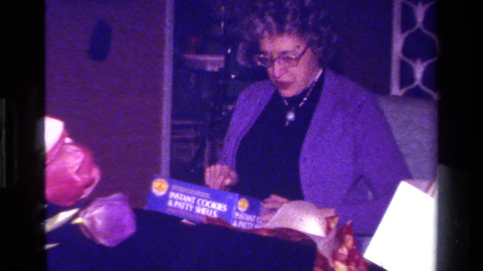 A woman in glasses and a purple jacket holds a box labeled as instant cookies and patty shells in a private interior.