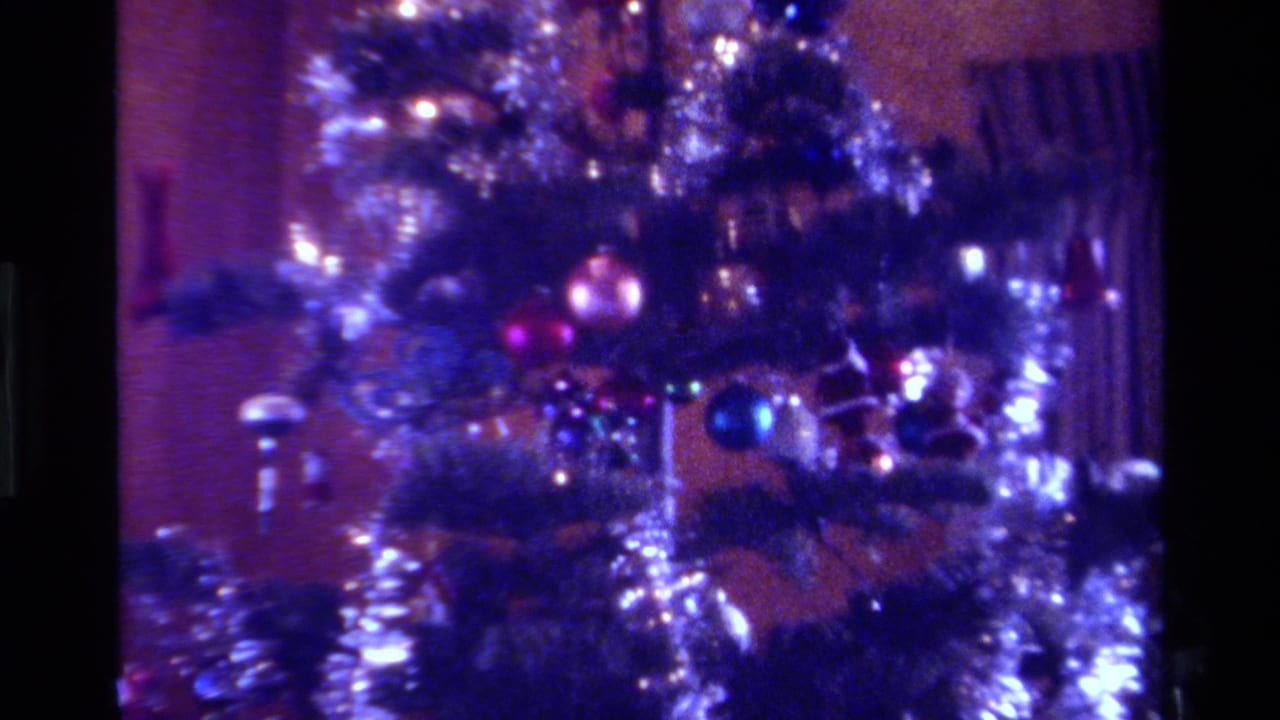 1979 California holiday home-movie frame with tinsel garland, blue and pink ornaments, and curtains behind the tree.