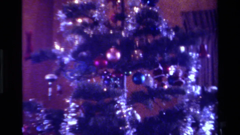 1979 California holiday home-movie frame with tinsel garland, blue and pink ornaments, and curtains behind the tree.