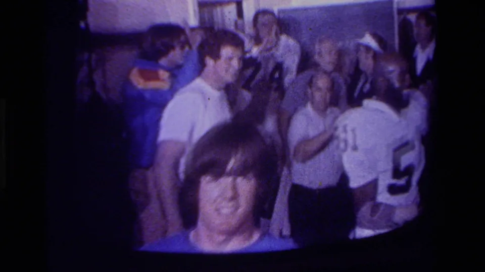 Indoor 8mm home-movie frame showing adults gathered around football players or fans in jerseys in California.