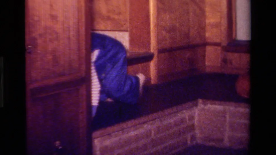 1970s California home-movie frame of a wood-paneled interior with a person partly visible behind furniture.